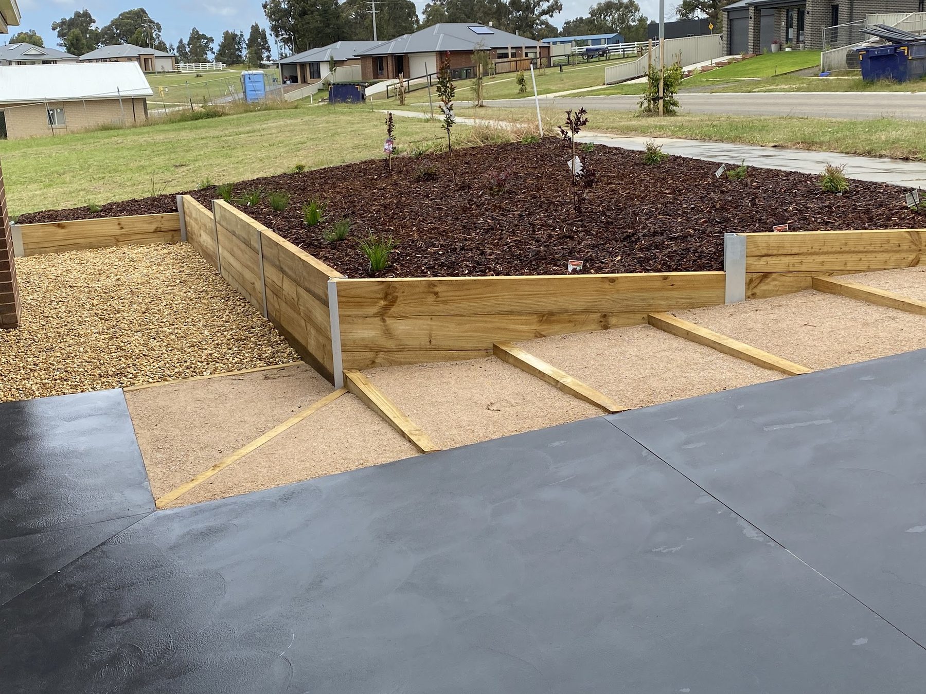 A stepped treated pine retaining wall with galvanised posts, holding a mulched garden bed above a decorative concrete driveway