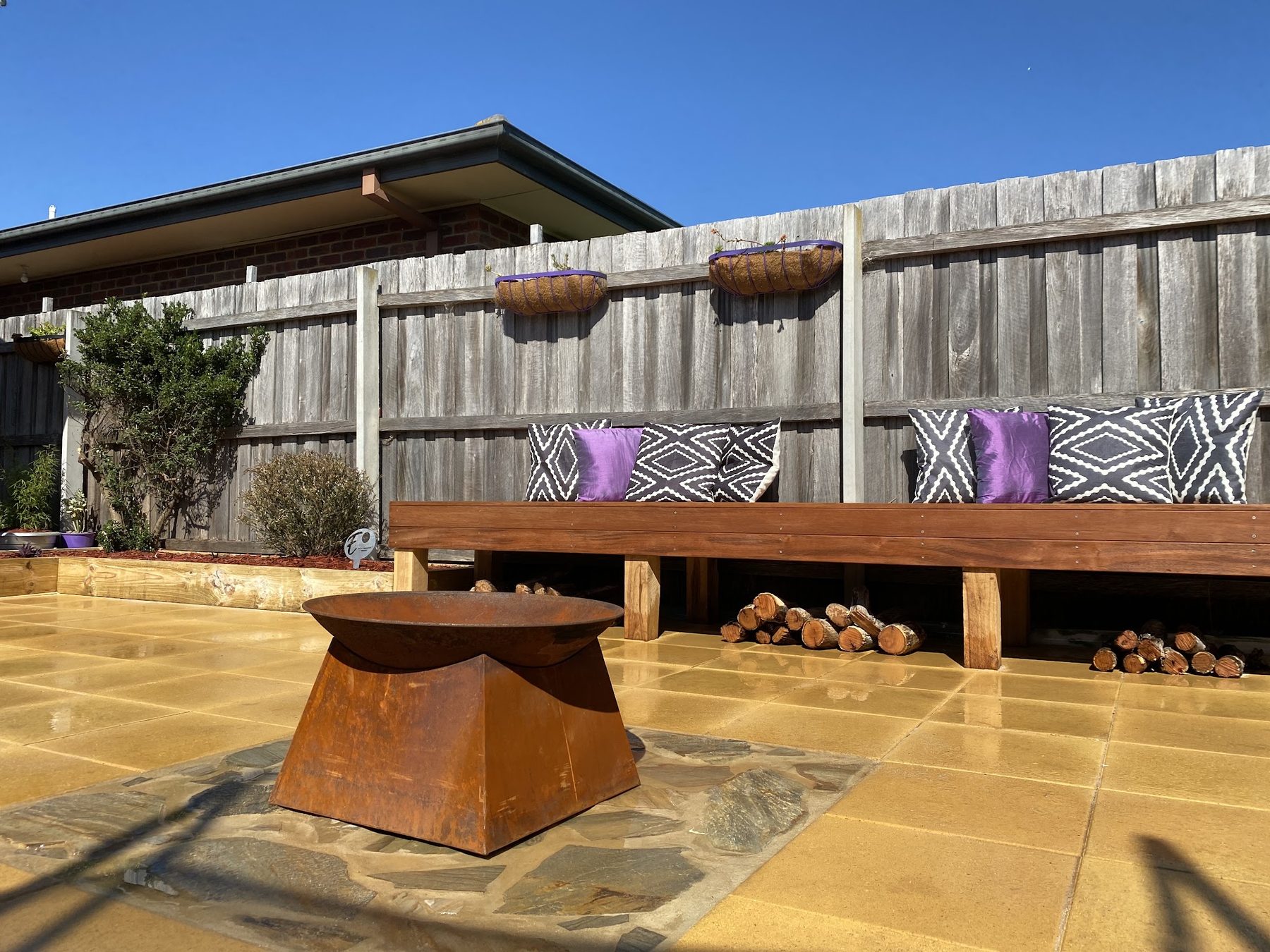 A paved courtyard built by Elliott’s Landscape and Design, with a fire bowl set into stone and a long built in bench along the fence