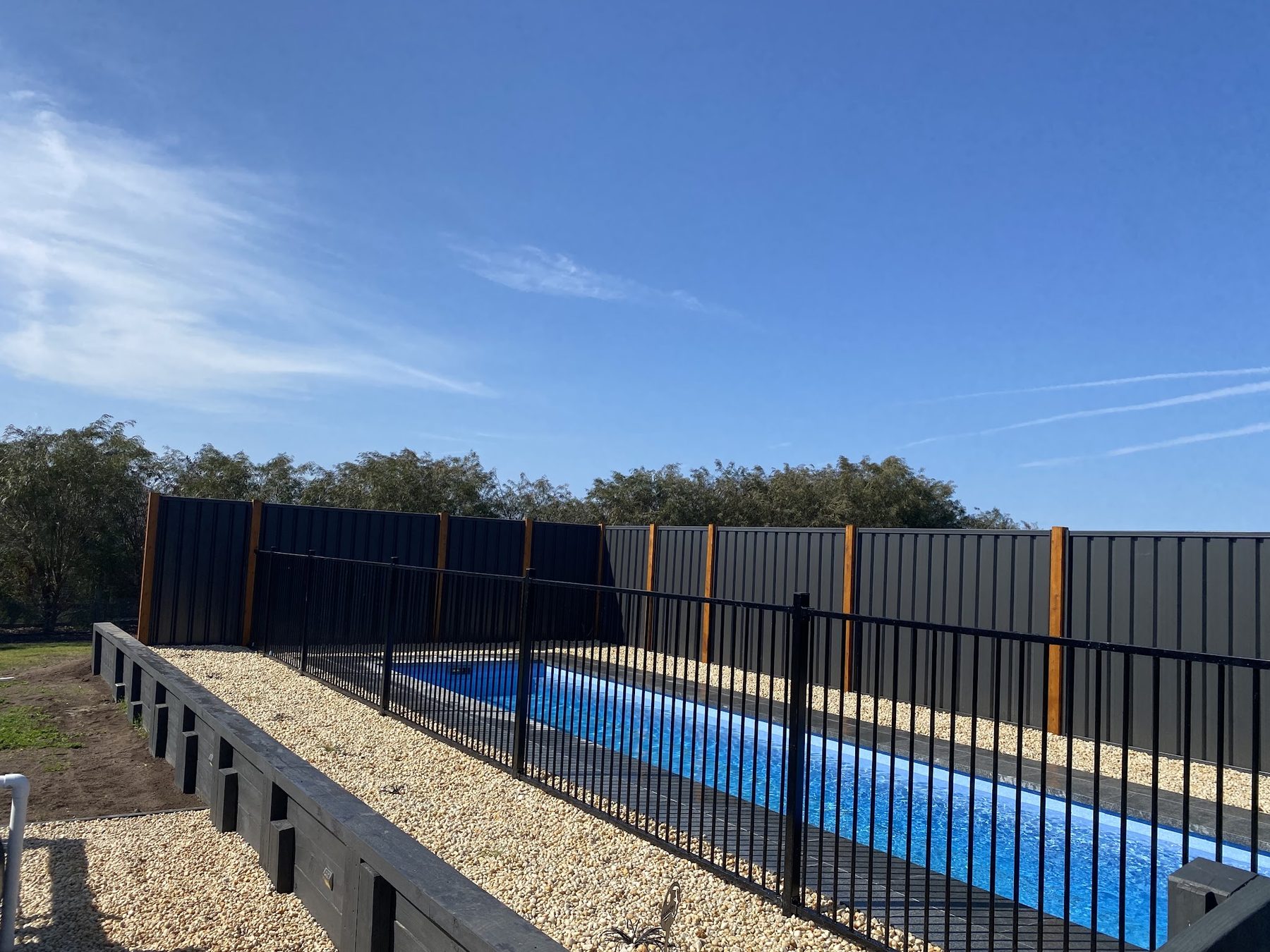 A pool surround with a pebble bed, sleeper retaining wall, black pool fence and new colorbond boundary fence